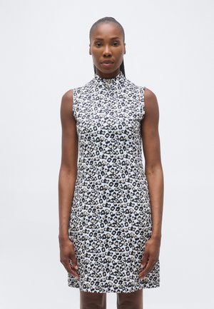 Kvinne som står med avslappede armer, iført ermeløs hvit kjole med svart og beige leopardmønster på ensfarget hvit bakgrunn.