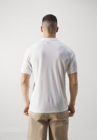 Polo blanc à manches courtes, tissu à gestion de l'humidité, avec une petite étiquette rouge "TÜRKİYE" au dos du col. Texture lisse.