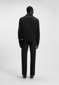 Veste bomber noire avec une taille et des poignets côtelés, portée sur un pantalon noir et des chaussures noires. Tissu lisse avec un design de col classique.