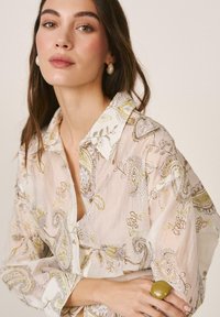 Mujer con cabello largo y castaño que lleva pendientes de perlas y una blusa ligera con estampado floral y bordados en verde y beige.