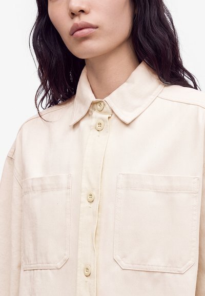 Femme aux cheveux ondulés foncés portant une chemise beige clair à boutons avec deux poches poitrine et un col pointu, montrée du cou à la poitrine.