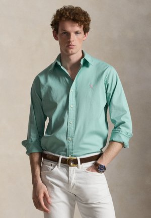 Joven con cabello rizado que lleva una camisa de botones verde claro, pantalones blancos, cinturón marrón y reloj de pulsera, de pie con una mano en el bolsillo.