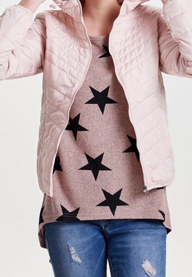 Personne portant une veste matelassée rose clair par-dessus une chemise beige avec des étoiles noires et un jean bleu déchiré sur un fond uni.