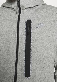 Šedá mikina na zip z měkké tkaniny, s texturovaným černým panelem s zipem a reflexním logem Nike na hrudi.