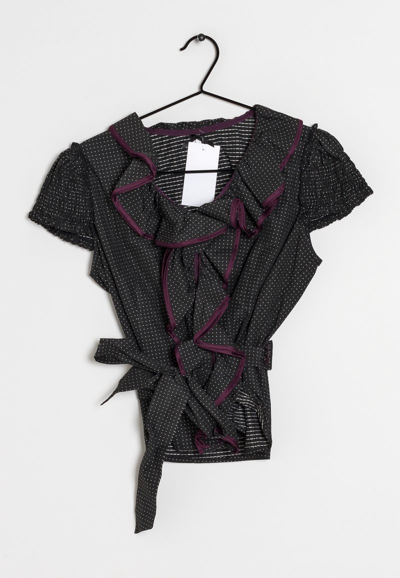 Haut cache-cœur noir avec manches courtes bouffantes ornées de pois blancs, bordure violette contrastante et volant à l'avant. Comprend un détail de lien en tissu.