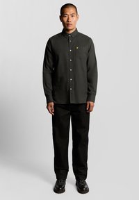 Camisa de botones de manga larga en verde oscuro con un bolsillo en el pecho y un logo amarillo. Combinada con pantalones negros y zapatos de cordones negros.