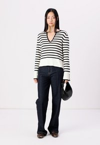 Striped black and white sweater with a V-neck, paired with dark blue flared jeans, and a black handbag. Slim fit with button accents.
