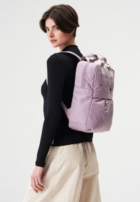 Lila Nylon-Rucksack mit doppelten oberen Griffen und verstellbaren Trägern, ausgestattet mit einer Fronttasche und minimalistischem Design. Glatte Textur.