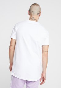 Weißes Baumwoll-T-Shirt mit kurzen Ärmeln, geradem Saum und lockerer Passform, getragen mit lila Jogginghosen. Sichtbare Tätowierungen an Armen und Hals.