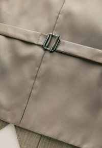 Beige stofjakke med en glat overflade, der har en sølvmetalknap og en skræddersyet design. Inkluderer en grøn stribet detalje nedenunder.