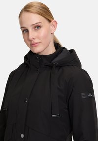 Schwarze isolierte Jacke mit Kapuze, vorderen Druckknöpfen, verstellbaren Kordeln und einer Logo-Patch am Ärmel. Glatte Textur.