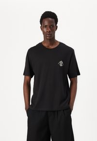 Black cotton t-shirt with short sleeves and a round neckline. Features a small, white sun and eye design on the left chest area.