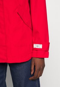 Chaqueta impermeable roja con un bolsillo, puños enrollados y una etiqueta. El material parece liso. Mano colocada en el detalle de la manga.