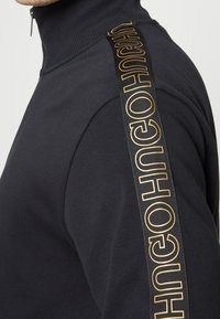 Sudadera negra con cremallera en el cuello y cuello acanalado. Presenta una franja dorada con el logo a lo largo del hombro. Tejido suave y liso, diseño ajustado.