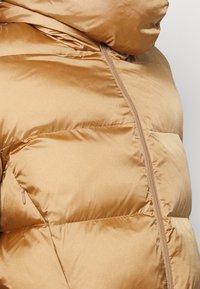 Giacca puffer marrone chiaro con colletto alto, texture trapuntata, zip frontale e dettagli delle tasche laterali. Materiale liscio e lucido.