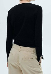 Personne portant un haut à manches longues noir et un pantalon beige avec des poches arrière, se tenant avec une main dans la poche contre un fond uni.