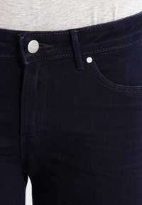 Mörkblå denimjeans har en klassisk femficksdesign, en silverknappstängning och kontrasterande sömmar. Tyget har en jämn textur.