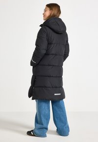 Schwarze Daunenjacke mit Kapuze, ausgestattet mit einem gesteppten Design, reflektierenden Akzenten und einem knielangen Schnitt. Kombiniert mit weit geschnittenen blauen Jeans.