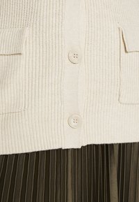 Krämfärgad stickad cardigan med ribbad textur, som har två framfickor och runda knappar. Nedan syns en plisserad mörk olivgrön kjol.