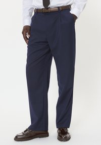 Granatowe spodnie garniturowe wykonane z gładkiego materiału, w prostym kroju z dostawem, z przodu zaszewkami i bocznymi kieszeniami, noszone z ciemnobrązowymi butami.
