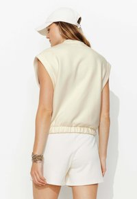 Haut sans manches beige, surdimensionné, avec une finition texturée, ourlet élastique, associé à un short blanc. Casquette blanche portée légèrement de biais.