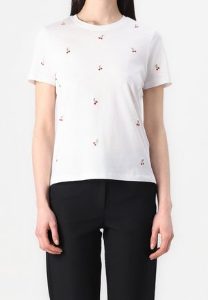 Kobieta ubrana w biały t-shirt z krótkim rękawem z małymi czerwonymi wzorami wiśni oraz czarne spodnie, pokazana od szyi do bioder.
