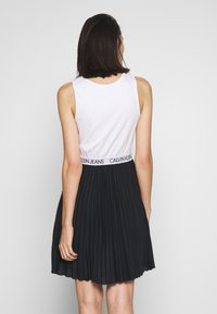 Vit ärmlös topp med en svart plisserad kjol. Midjebandet har en "CALVIN JEANS"-logotyp i svart på vit bakgrund. Slät och texturerad tyg.