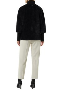 Vrouw in een zwart, gestructureerd, kort jasje, witte, op maat gemaakte broek en zwarte, hooggehakte enkellaarsjes, staand met haar rug naar de camera op een witte achtergrond.