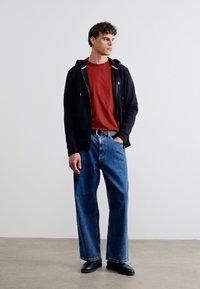 Νέος άνδρας στέκεται φορώντας μαύρο φούτερ με φερμουάρ, κόκκινη μπλούζα, φαρδιά μπλε τζιν και μαύρα παπούτσια, κοιτάζοντας δεξιά σε λευκό φόντο.