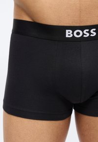 Gros plan sur une personne portant un boxer noir avec une large ceinture arborant le logo blanc "BOSS" à l'avant.