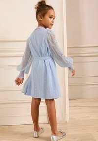 Vestito azzurro chiaro con tessuto trasparente, texture a pois, dettagli floreali ricamati sulle maniche e vita con cintura. Ballerine argentate completano il look.