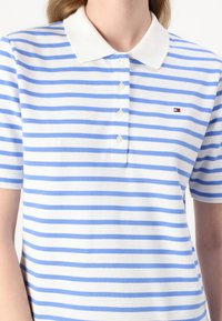 Stripete polo skjorte i lyseblå og hvit, laget av mykt stoff. Har en hvit krage og tre knapper, med en liten logo på brystet.