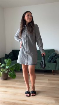 Nő szürke gombos ingruhában, fekete fonott kézitáskát tartva, egy nappaliban áll, ahol zöld kanapé és cserepes növény található.