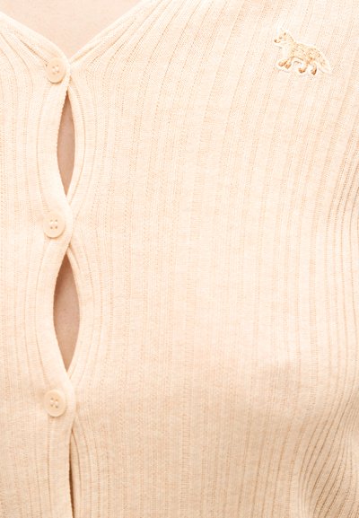 Μπεζ ριμπ πλεκτή ζακέτα με τρία μπεζ κουμπιά και μικρό κεντημένο λογότυπο αλεπούς στην επάνω αριστερή πλευρά.