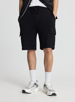 Mężczyzna ubrany w czarne szorty typu cargo, białe skarpetki oraz białe i szare buty sportowe, stoi z rękami w kieszeniach na tle jednokolorowym.