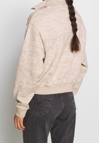 Beige collegepaita, jossa on hienovarainen tekstuuri, korkea kaulus ja resorit hihansuissa, yhdistetty tummanharmaisiin farkkuihin, joissa on sydämenmuotoiset takataskut.