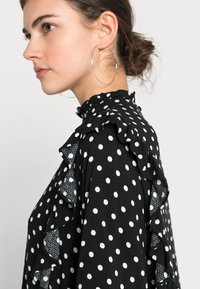 Blusa preta com bolinhas brancas, apresentando um decote e mangas ondulados. Textura suave, tecido leve, design ajustado.