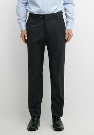 Uomo con pantaloni eleganti neri, scarpe eleganti in pelle nere e camicia azzurra a righe con bottoni, in piedi su uno sfondo bianco semplice.