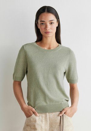 Frau mit glatten dunklen Haaren, die einen hellgrünen kurzärmligen Strickpullover und beige Hosen trägt, steht mit den Händen in den Taschen vor einer weißen Wand.