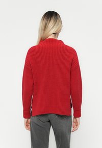 Roter Strickpullover mit hohem Kragen, überschnittenen Schultern und gerippten Bündchen. Seitliche Schlitze am Saum. Kombiniert mit grauen Hosen.