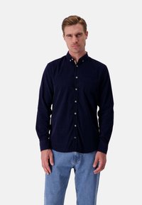 Camicia blu navy con colletto, dotata di tasca frontale e maniche lunghe. Il tessuto appare liscio con un aspetto texturizzato. Abbinata a jeans azzurri chiari.