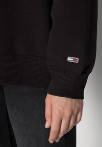Primo piano di una mano accanto al polsino rigato di un felpa nera con un piccolo logo rettangolare ricamato in rosso, bianco e blu.