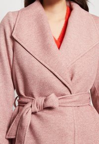 Donna che indossa un morbido cappotto di lana rosa con cintura sopra un maglione V-incrociato arancione brillante e peloso, mostrata dalla collana alla vita.