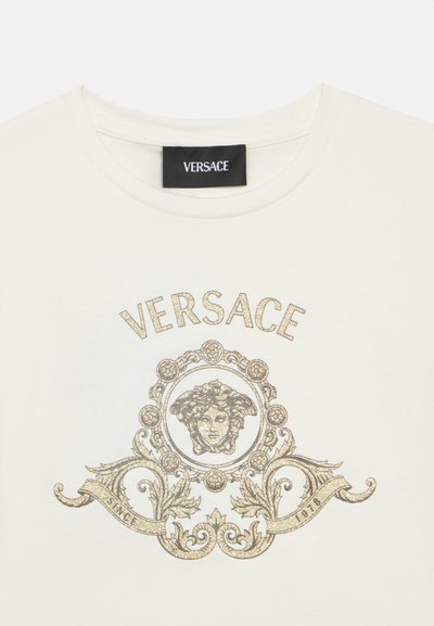 Λευκό t-shirt Versace με χρυσό λαμπερό έμβλημα κεφαλής Μέδουσας και περίτεχνο λουλουδάτο σχέδιο, κείμενο "VERSACE" πάνω και "Since 1978" σε λωρίδα από κάτω.