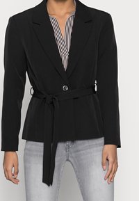 Zwarte blazer met een aansluitend ontwerp, lange mouwen, een knoopsluiting en een tailleriem. Draagt men over een zwart-wit gestreepte top.