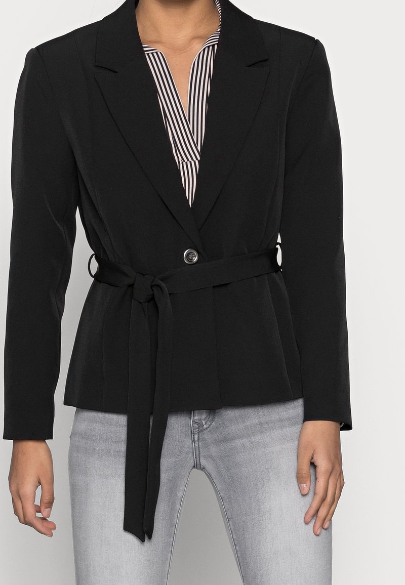 Zwarte blazer met een aansluitend ontwerp, lange mouwen, een knoopsluiting en een tailleriem. Draagt men over een zwart-wit gestreepte top.