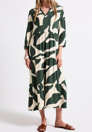 Kvinde iført en løs midikjole med lange ærmer og V-hals med grønne og beige abstrakte mønstre og brune flade sandaler.
