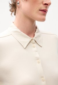 Beige Hemd mit Kragen und glatter Textur, mit einer Knopfschließung an der Vorderseite und einem subtilen Glanz.