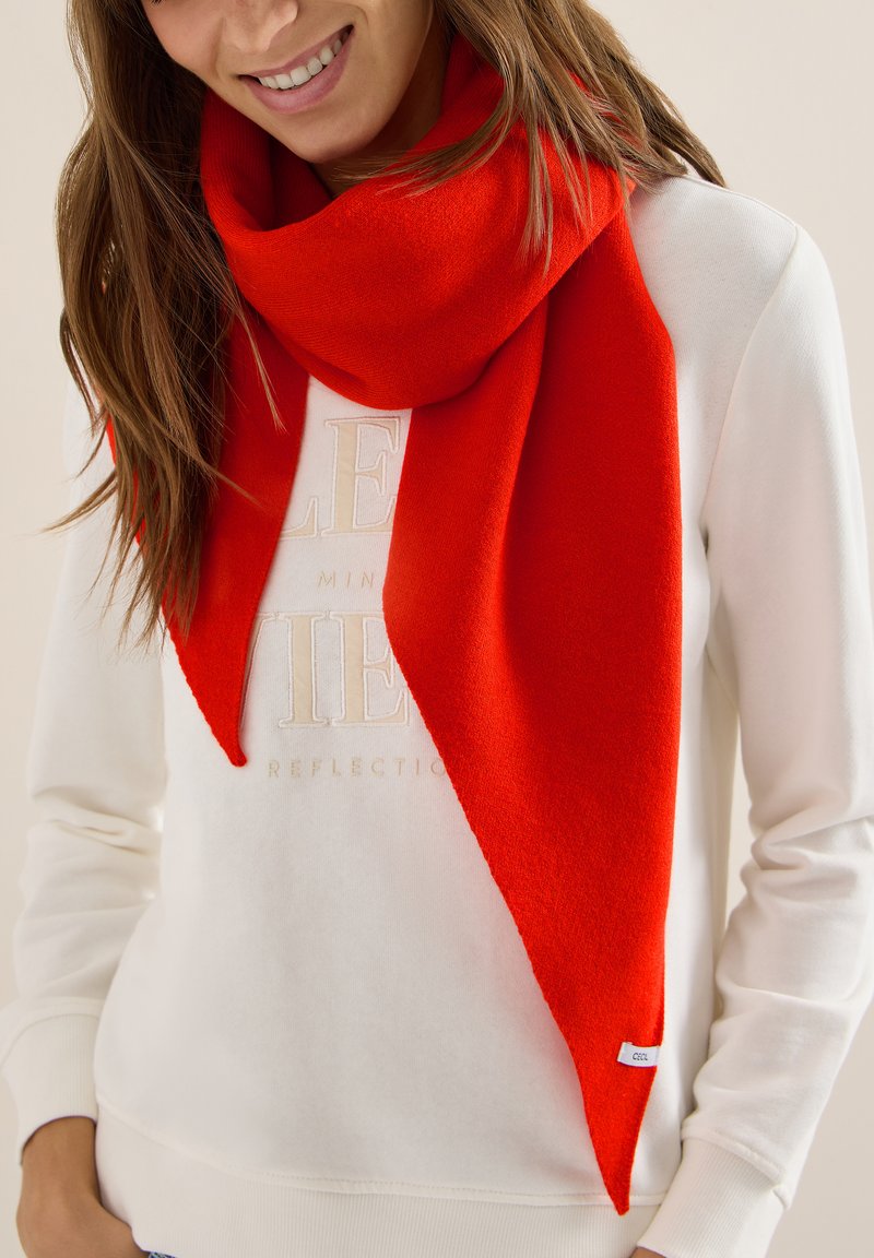 Femme souriante portant une écharpe rouge vif sur un sweat-shirt blanc à manches longues avec un texte brodé subtil.
