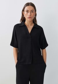 Zwarte shirt met korte mouwen en een kraag, lichtgewicht textuur en een relaxte pasvorm. Te combineren met bijpassende zwarte broek.